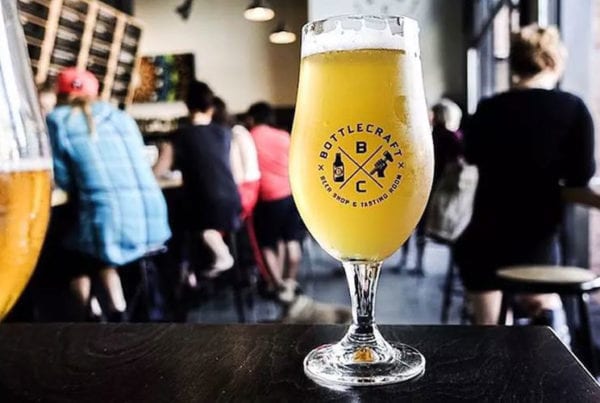 A close up of a pint of beer with the Bottlecraft logo. Patrons sit in the background.