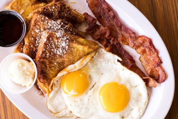 An overhead view of two eggs, french toast, two strips of bacon, syrup and butter.